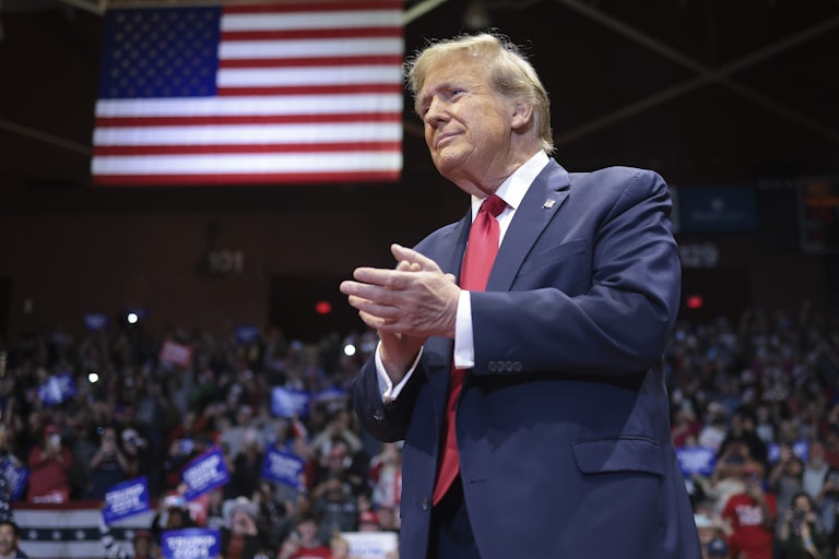 Donald Trump claps. A crowd is in the background, and a large U.S. flag hangs from the ceiling.