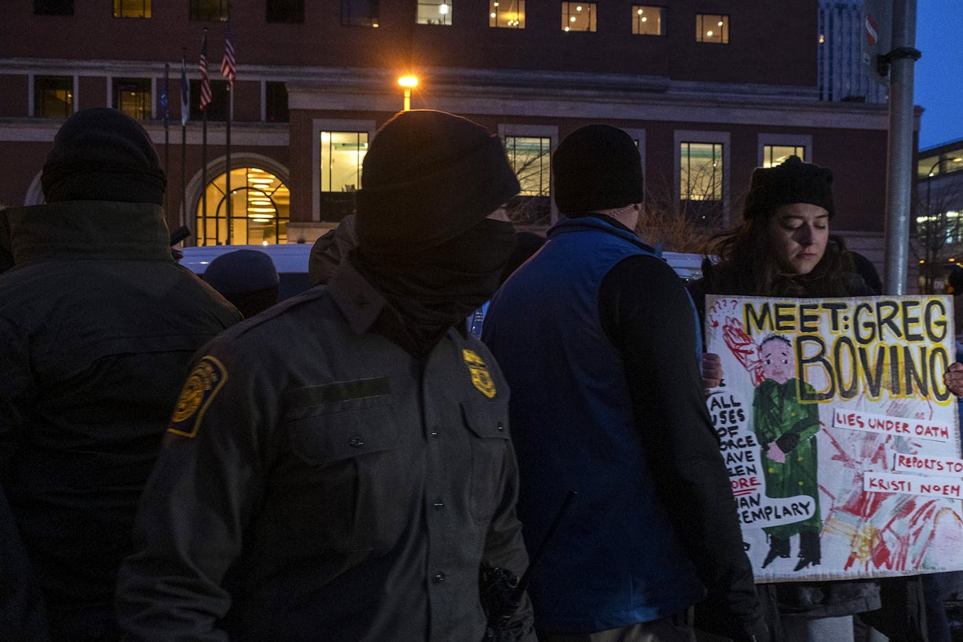 A protester holds a poster that says "Meet: Greg Bovino. Lies under oath. Reports to Kristi Noem. All uses of force have been more than exemplary."