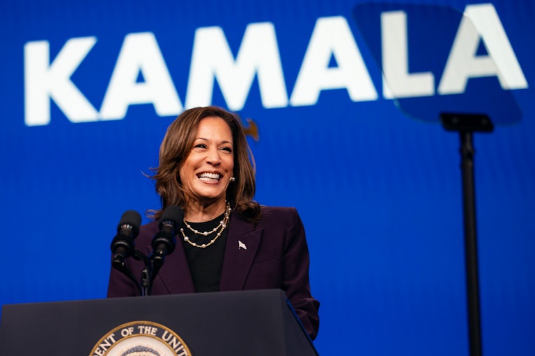 Kamala Harris smiles at a podium. Behind her is a blue backdrop with "KAMALA" in giant letters.