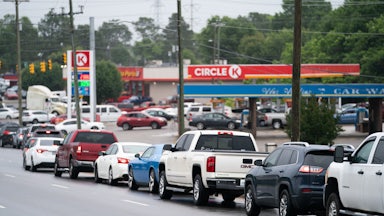 Cars on the road wait in line to drive into a gas station.
