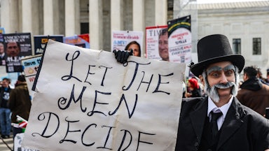 A demonstrator in historical costume holds a sign reading "let the men decide"