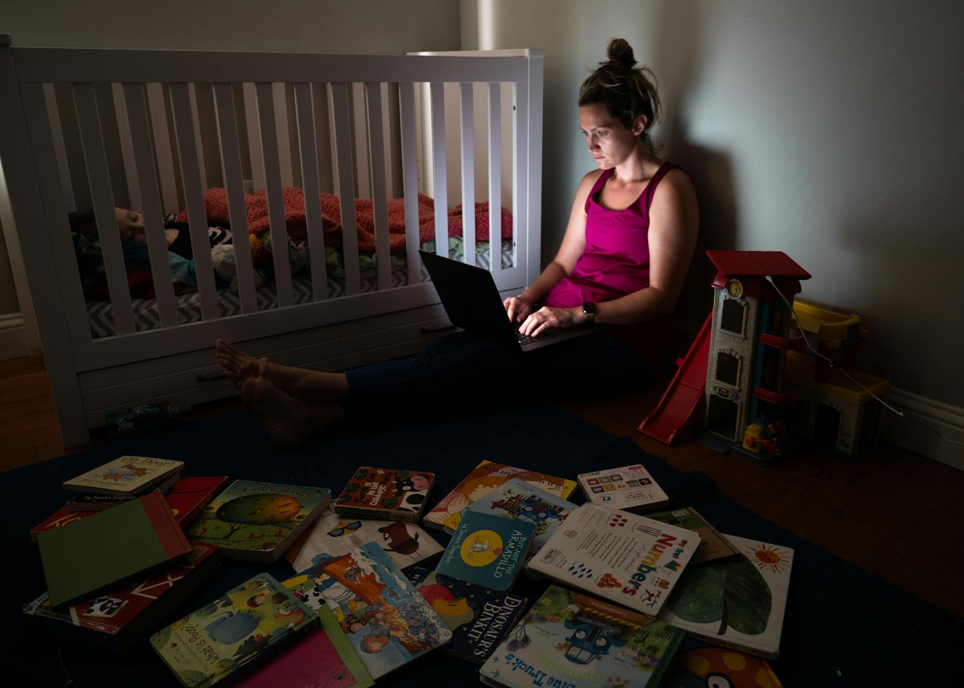 A tired mom works on her laptop next to a crib in a cluttered nursery. The title is Manages Time Effectively from the series “Résumé of a Stay at Home Mom” 