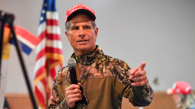 Dan Meuser wears camo while standing in front of an American flag holding a microphone and gesticulating.