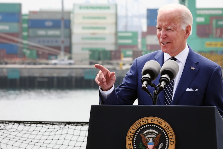 President Biden speaks at a podium, with a harbor and shipping containers visible behind him.