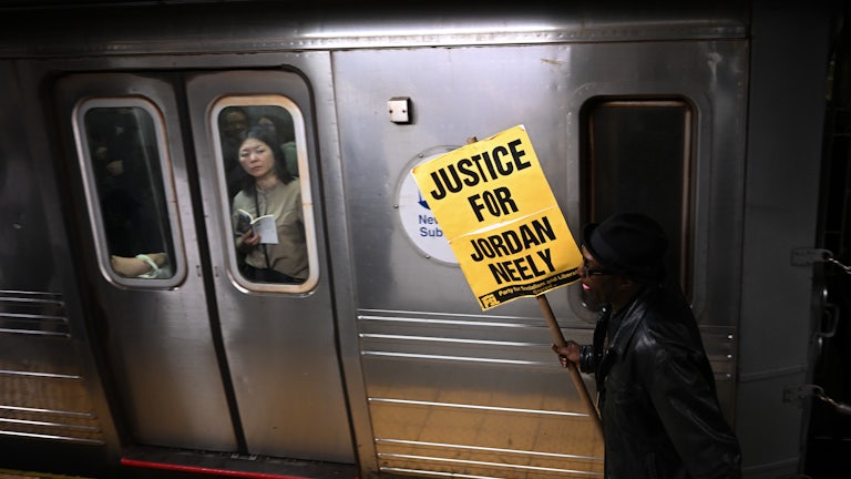 A woman riding the subway looks at a protestor carrying a “Justice for Jordan Neely” poster walking on the platform.