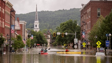 People kayak down a flooded city street; wooded mountains rise up in the background. 
