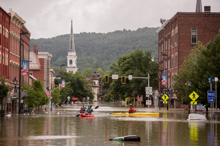 People kayak down a flooded city street; wooded mountains rise up in the background. 