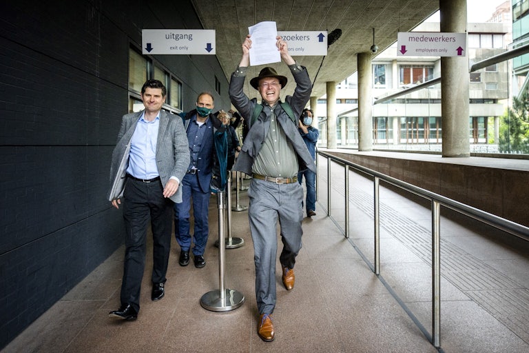 A man grins holding a piece of paper over his head triumphantly as he walks. To his left, another man smiles. Others follow.