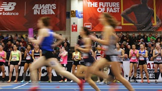Three young women runners run by, blurred, on an indoor track, as other young runners and spectators look on.