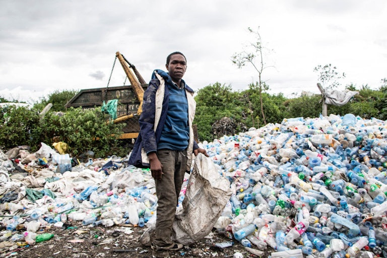 A man holding a bag stands in front of a large mound of plastic trash.