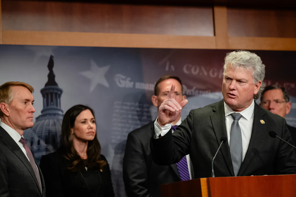 Mike Collins gesticulates while standing at a lectern, with others in suits behind him.
