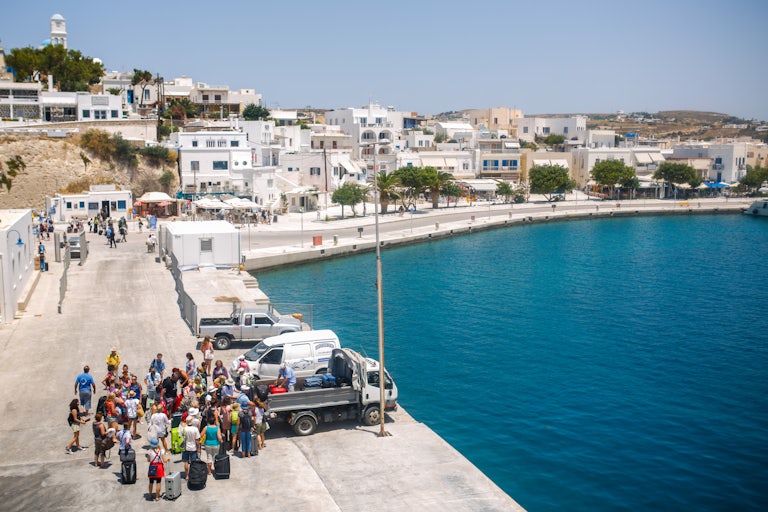 A view from above of a harbor with white buildings set against a deep blue sea. A throng of people with suitcases are gathered in the bottom left of the photo, taking bags off a truck.