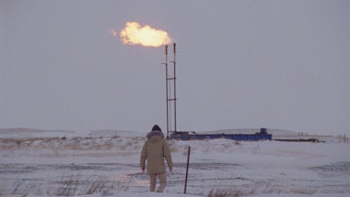 A person walks across a frozen field with a gas or oil flare in the background.
