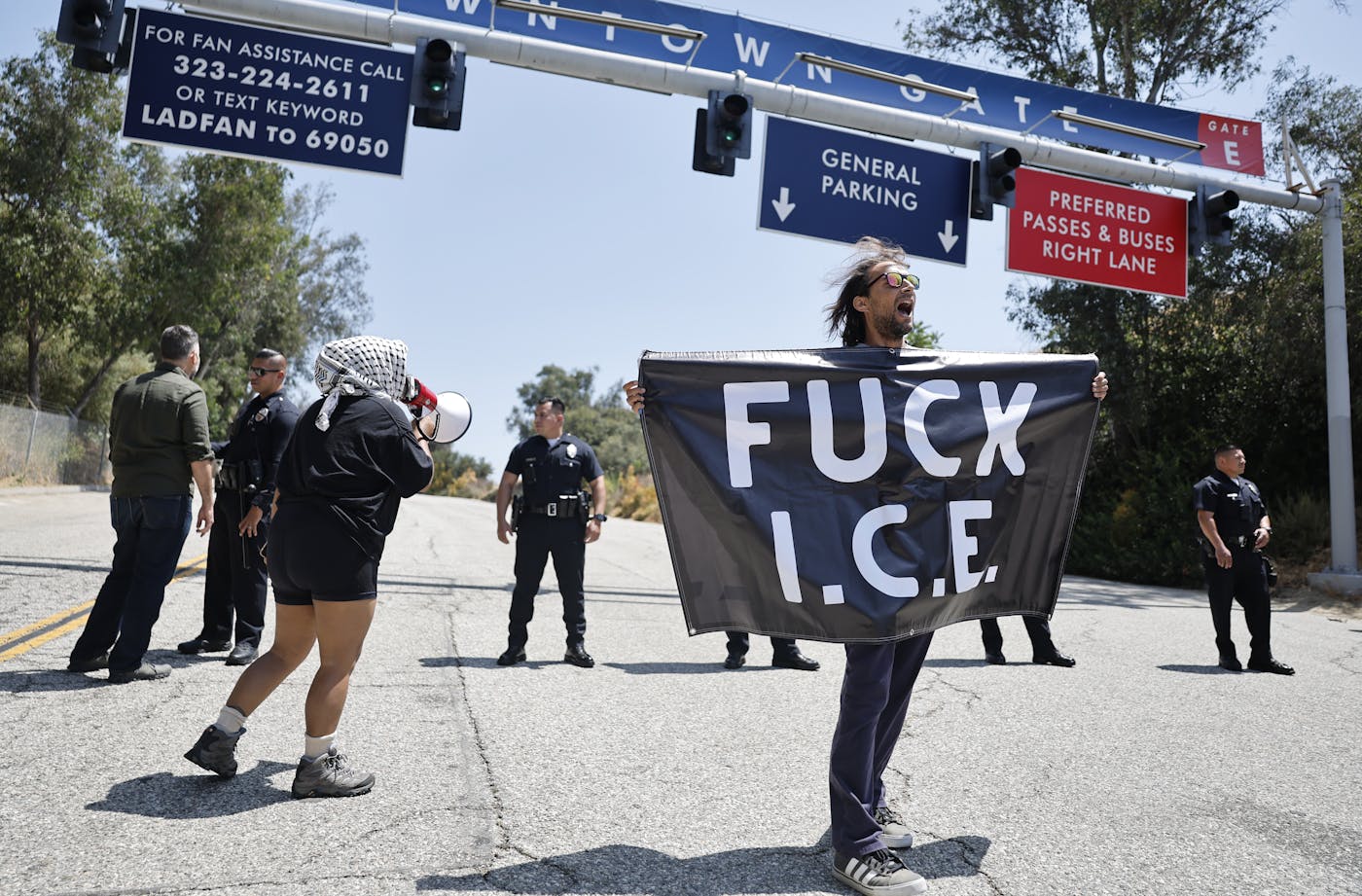 A man unfurls a banner that reads âFUCK I.C.E.â Someone with a keffiyeh around their head yells at police officers stationed outside Dodgers Stadium.
