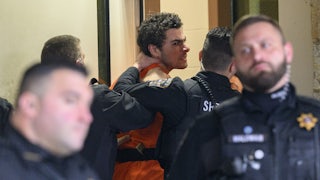 Luigi Mangione wearing an orange jumpsuit makes an angry face as police officers push him, one with a hand on his neck