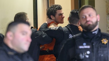 Luigi Mangione wearing an orange jumpsuit makes an angry face as police officers push him, one with a hand on his neck