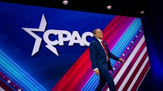 Ken Paxton walks across a red, white, and blue stage with the CPAC logo behind him.