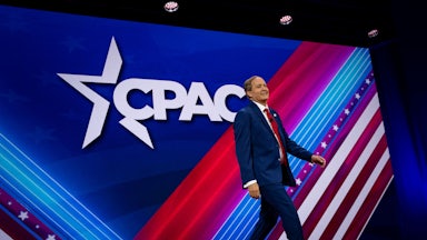 Ken Paxton walks across a red, white, and blue stage with the CPAC logo behind him.