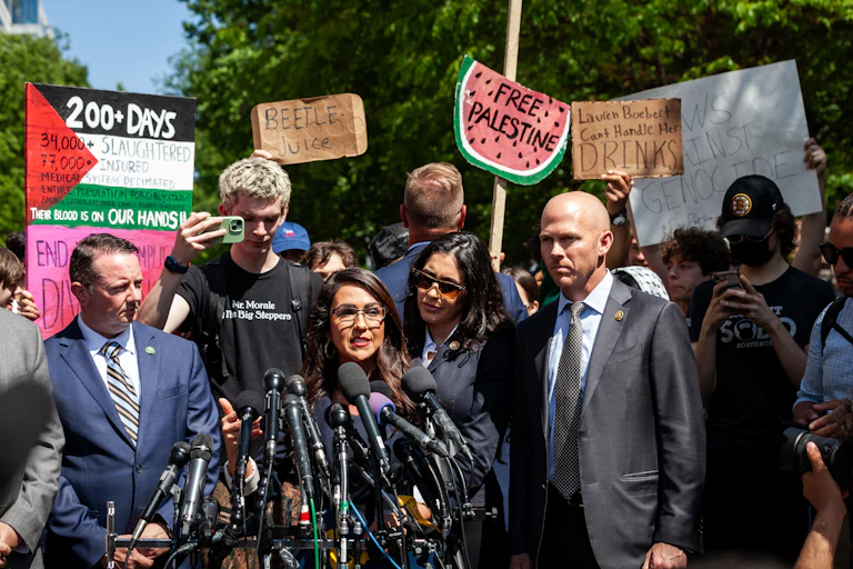 Lauren Boebert in a crowd. Signs behind her read "Beetlejuice," "Lauren Boebert can't handle her drinks," a watermelon that says "Free Palestine," and a Palestinian Flag that reads "200+ Days."