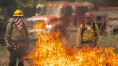 Two people in firefighting gear stand in front of a truck, with flames in front of them.