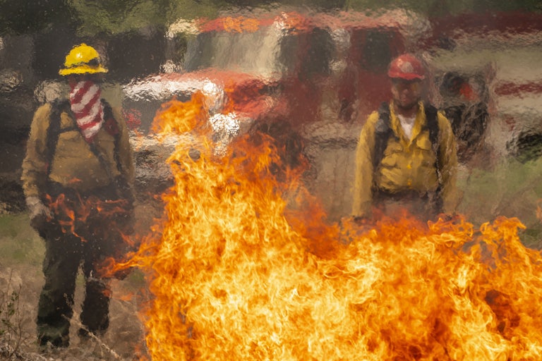 Two people in firefighting gear stand in front of a truck, with flames in front of them.