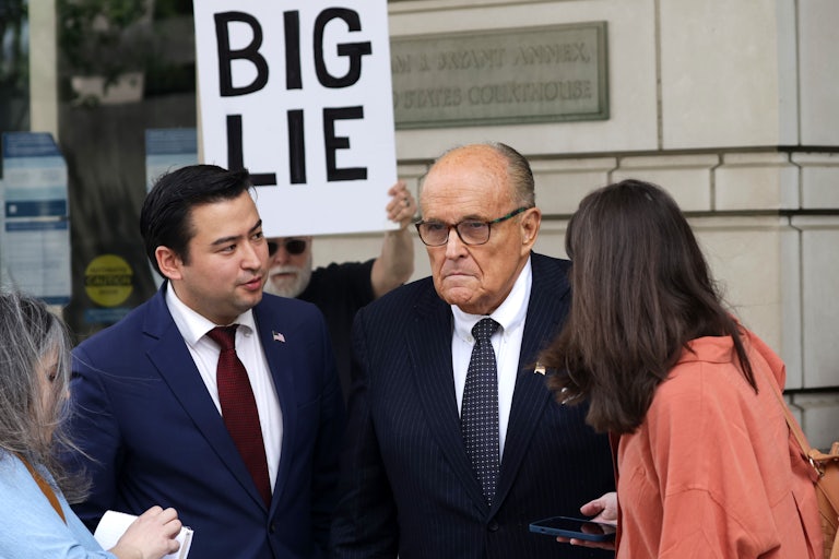 Rudy Giuliani frowns while someone behind him holds a poster that reads "Big Lie" in all caps