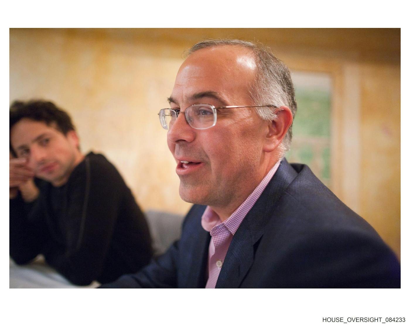New York Times columnist David Brooks speaks, smiling, at a table with others. One man, the only one pictured, looks over at him.