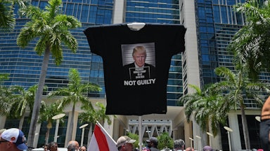 A man holds up a stick with a t-shirt attached to the top that says NOT GUILTY under a photo of Trump.