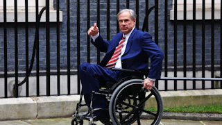 Greg Abbott rolls his wheelchair forward on the sidewalk and makes a thumbs up sign. A black fence is behind him.