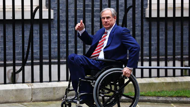 Greg Abbott rolls his wheelchair forward on the sidewalk and makes a thumbs up sign. A black fence is behind him.