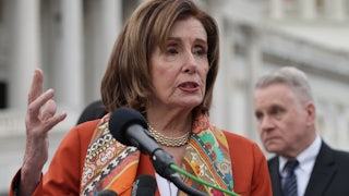 Nancy Pelosi speaks at a podium in front of the Capitol building while wearing an orange blazer and colorful scarf.