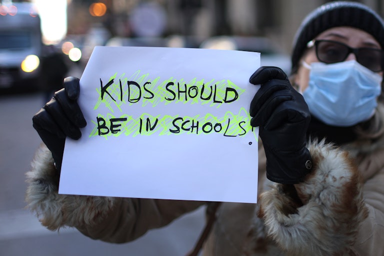 a masked person holding a sign that reads "kids should be in schools"