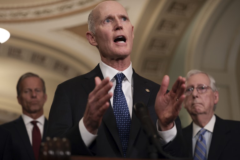 Florida Senator Rick Scott gestures with his hands at a press conference, flanked by John Thune and Mitch McConnell