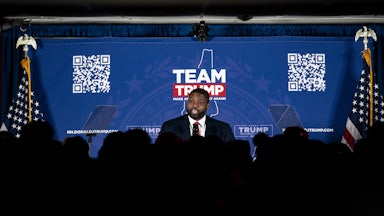 Byron Donalds stands on a stage in front of a background that says "Team Trump" and has a QR code. A crowd's silhouette is seen in front of him.