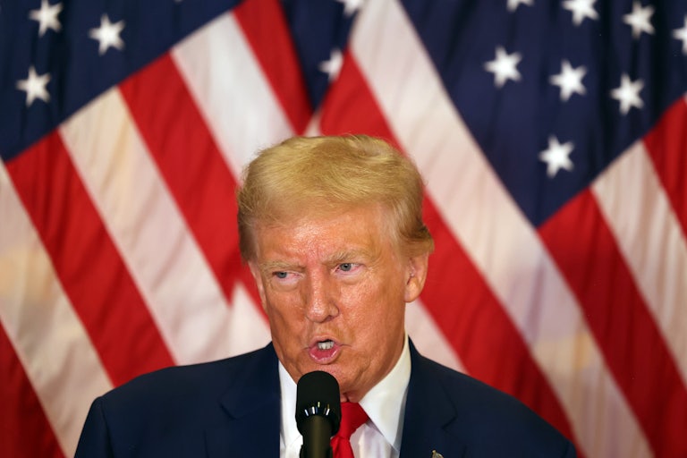 Donald Trump speaks at a mic. Several U.S. flags are behind him.
