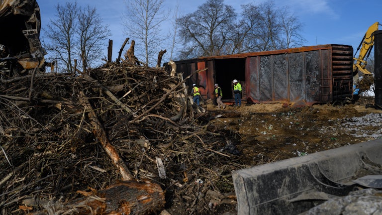 A cargo train, a giant pile of branches/uprooted trees, and three workers in the background.