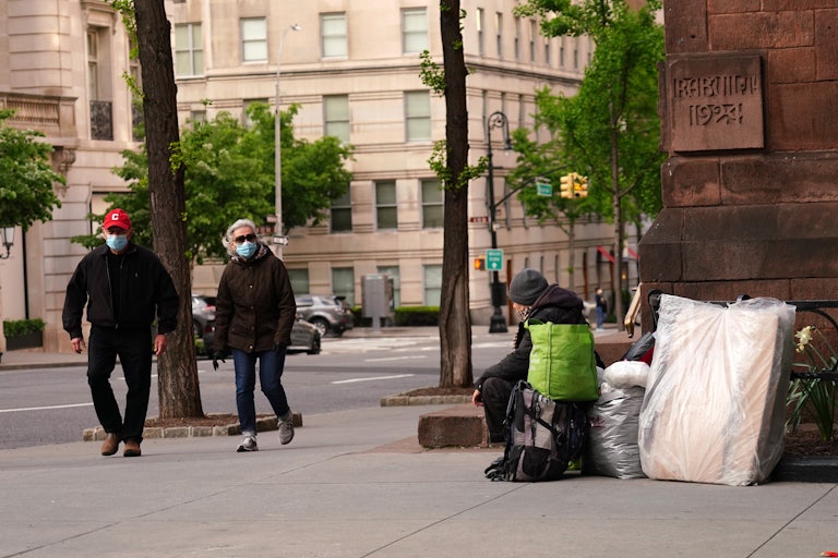 An unhoused person sitting on the sidewalk in Manhattan as pedestrians pass.