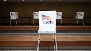 A lone voting booth stands in the foreground at a polling location.