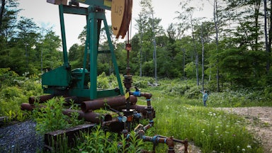 A pumpjack stands in a forest clearing, with a person in the background.