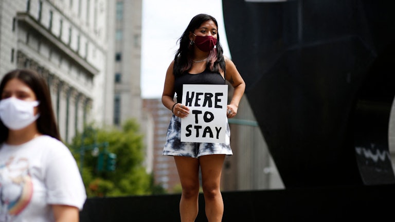A young woman holds a banner reading “Here to Stay”