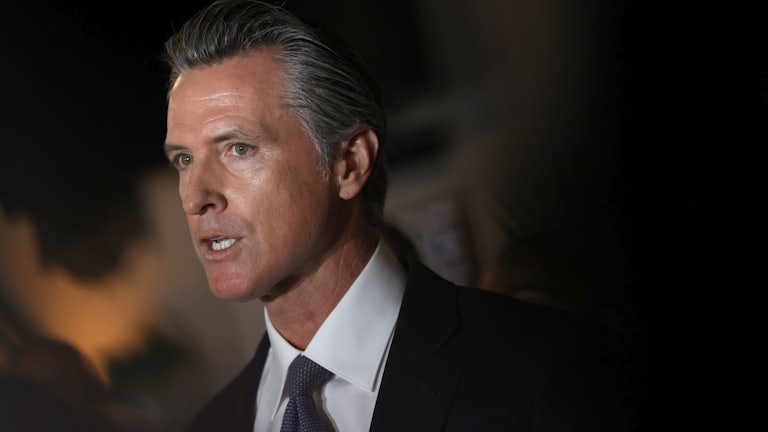 A close-up of Gavin Newsom standing in shadows as he speaks at a press conference.