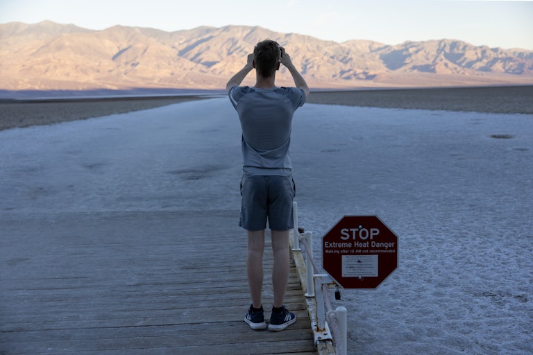 A shot of a man from the back, standing on a wooden platform taking a photo of the desert and mountains behind it.  Next to him, a sign says STOP: EXTREME HEAT DANGER.