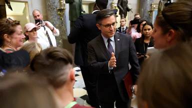 Mike Johnson stands in a group of people and gestures while talking.