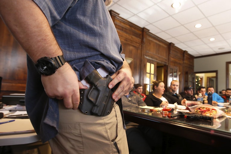 A man has a gun in the holster on his hap. His hands are on the gun as his shirt is tucked up.