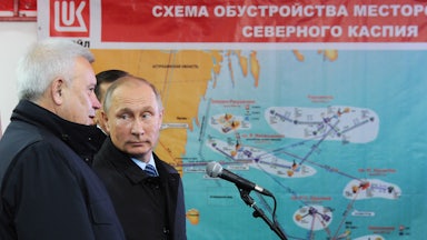 Vladimir Putin stands in front of an oil schematic while listening to another man talk.