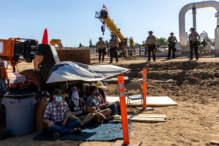 Activists sit beneath a tarp as law enforcement officers line up in front of construction equipment.