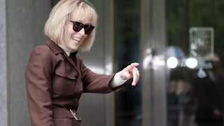 E. Jean Carroll smiles and points to something off camera. She's wearing a brown coat and sunglasses.