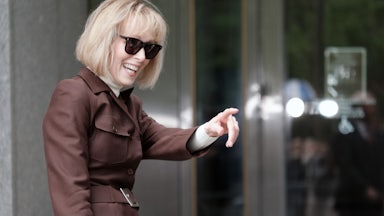 E. Jean Carroll smiles and points to something off camera. She's wearing a brown coat and sunglasses.