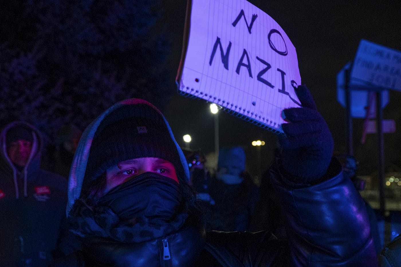 A man at an outdoor protest holds a sprial notebook with a page open that reads NO NAZIS in black marker