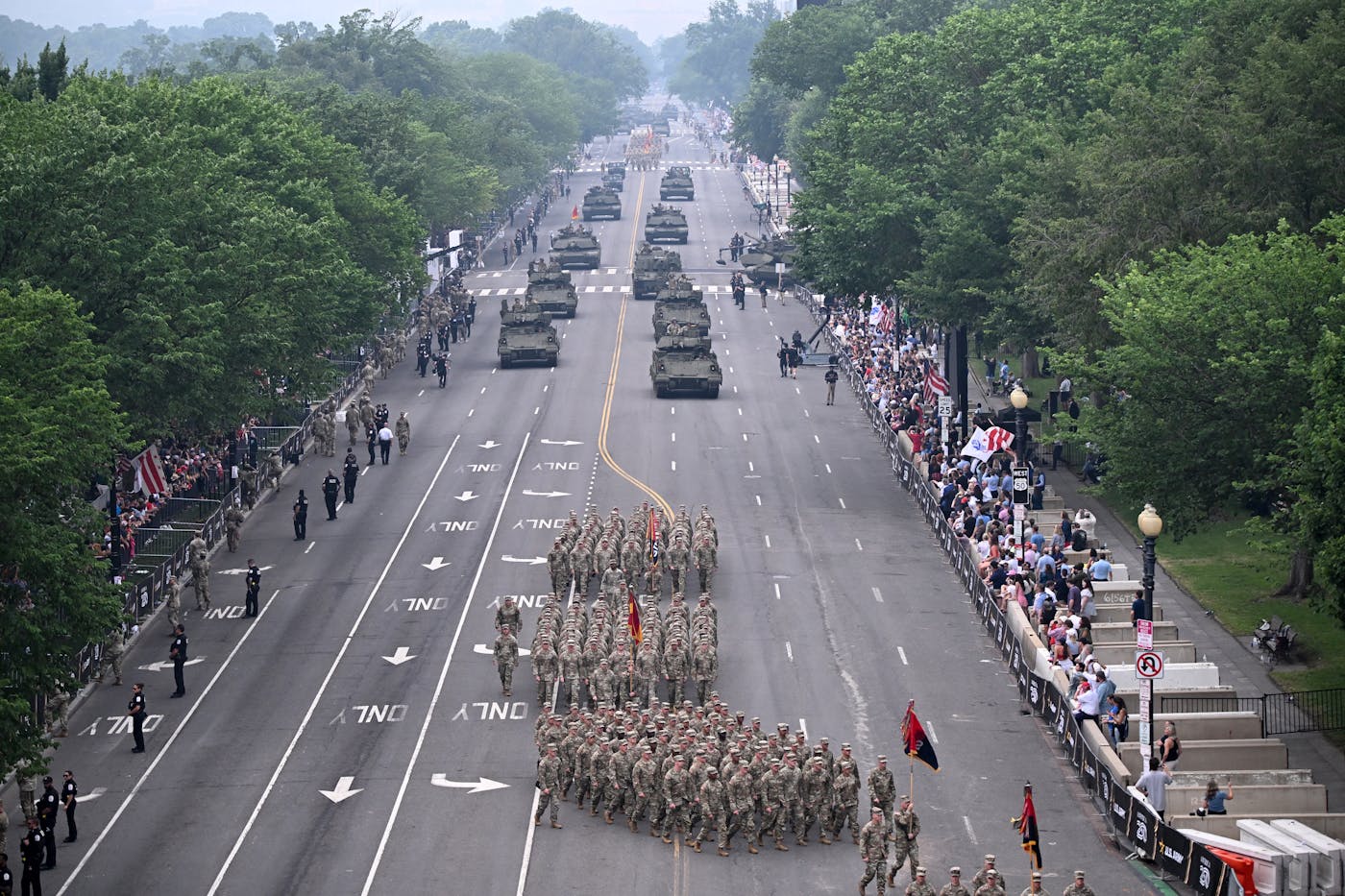 Army soldiers march and tanks roll down a street in Washington, D.C. as people stand on the streets watching them. It is not a large crowd.